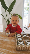 Child in a red shirt that says L is for Love, playing with a tic-tac-toe game on a wooden table.