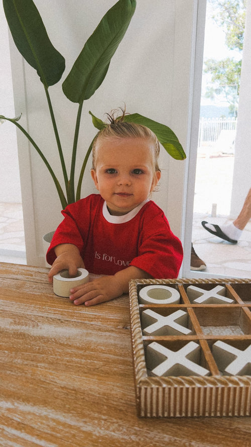 Child in a red shirt that says L is for Love, playing with a tic-tac-toe game on a wooden table.