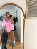 Person taking a mirror selfie wearing a pink oversized t-shirt with text, black shorts, and gray sneakers.