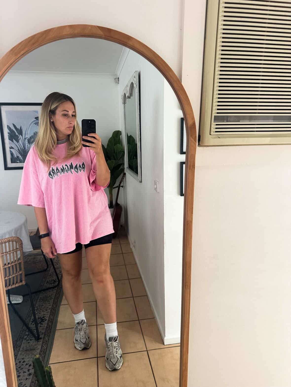 Person taking a mirror selfie wearing a pink oversized t-shirt with text, black shorts, and gray sneakers.