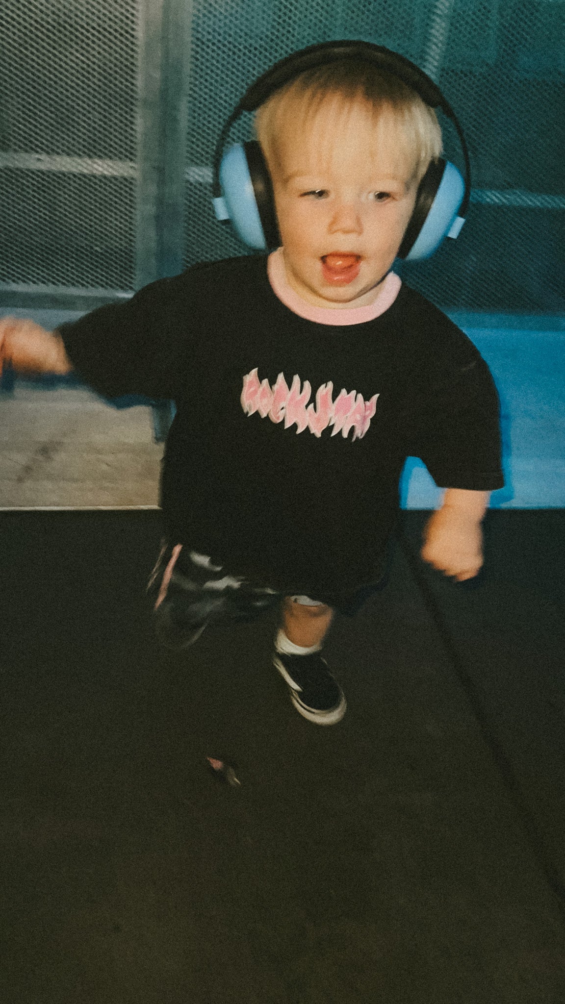 Child wearing headphones and a black t-shirt with pink text, standing on a dark surface.