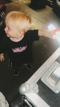 Child wearing a black t-shirt with pink text, standing next to a suitcase.