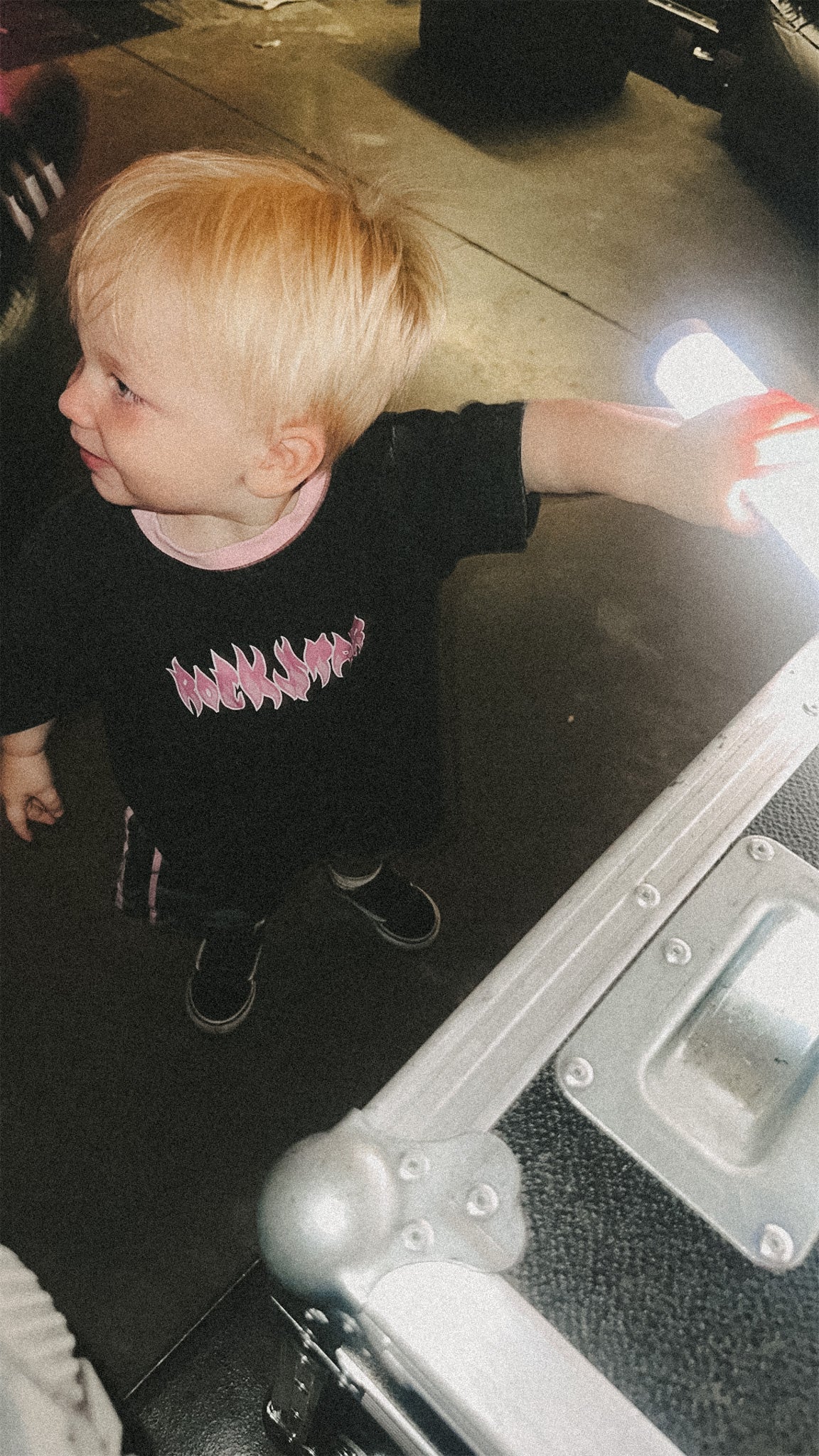 Child wearing a black t-shirt with pink text, standing next to a suitcase.