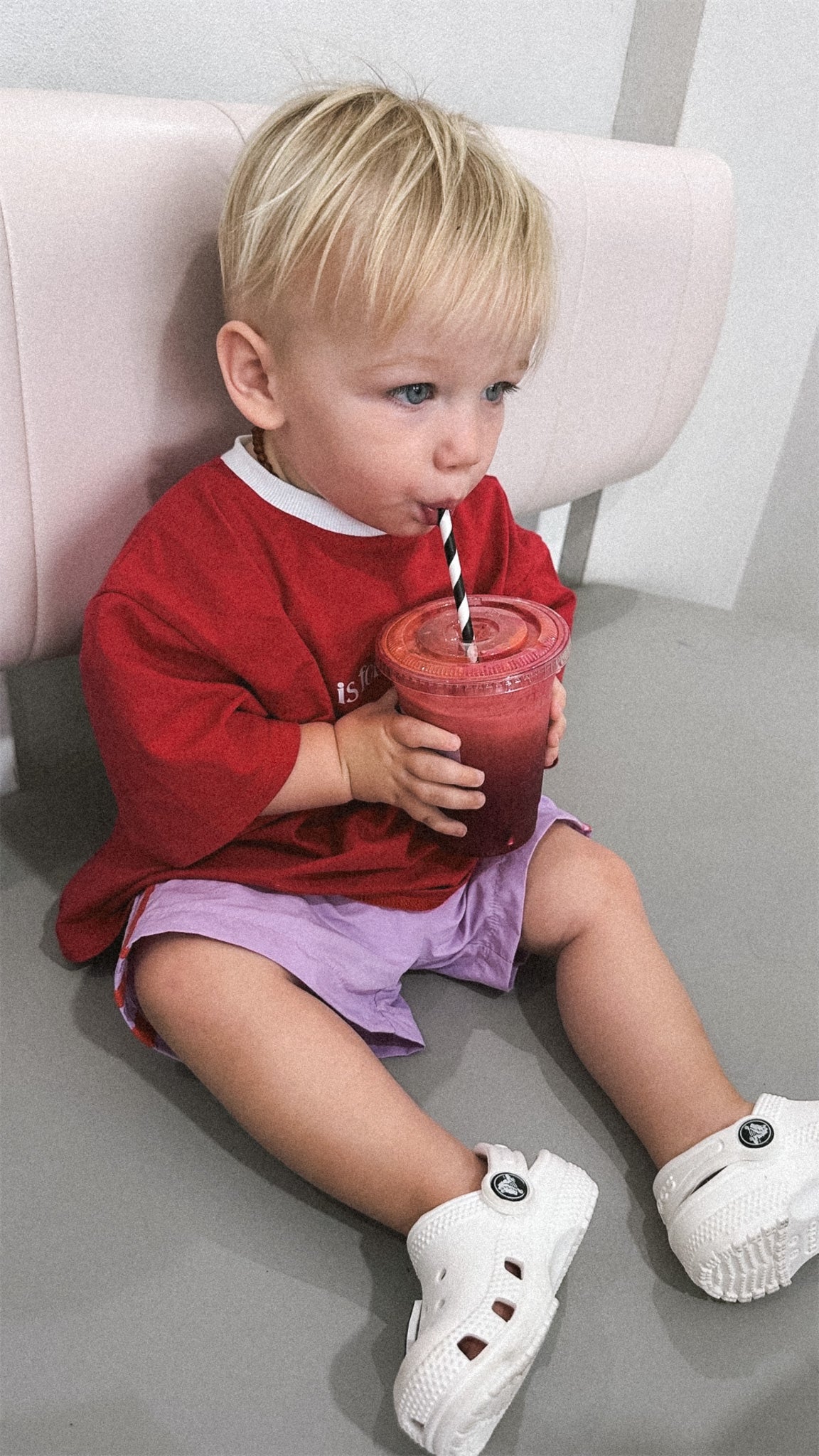 Child in a red shirt and purple shorts sitting on a couch, holding a red cup with a straw.