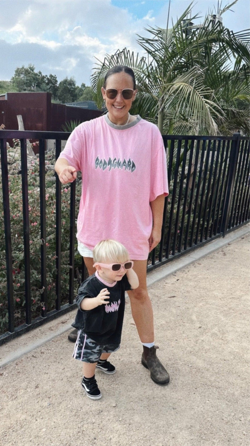 Woman in pink oversized t-shirt and sunglasses standing with a child in sunglasses outdoors.