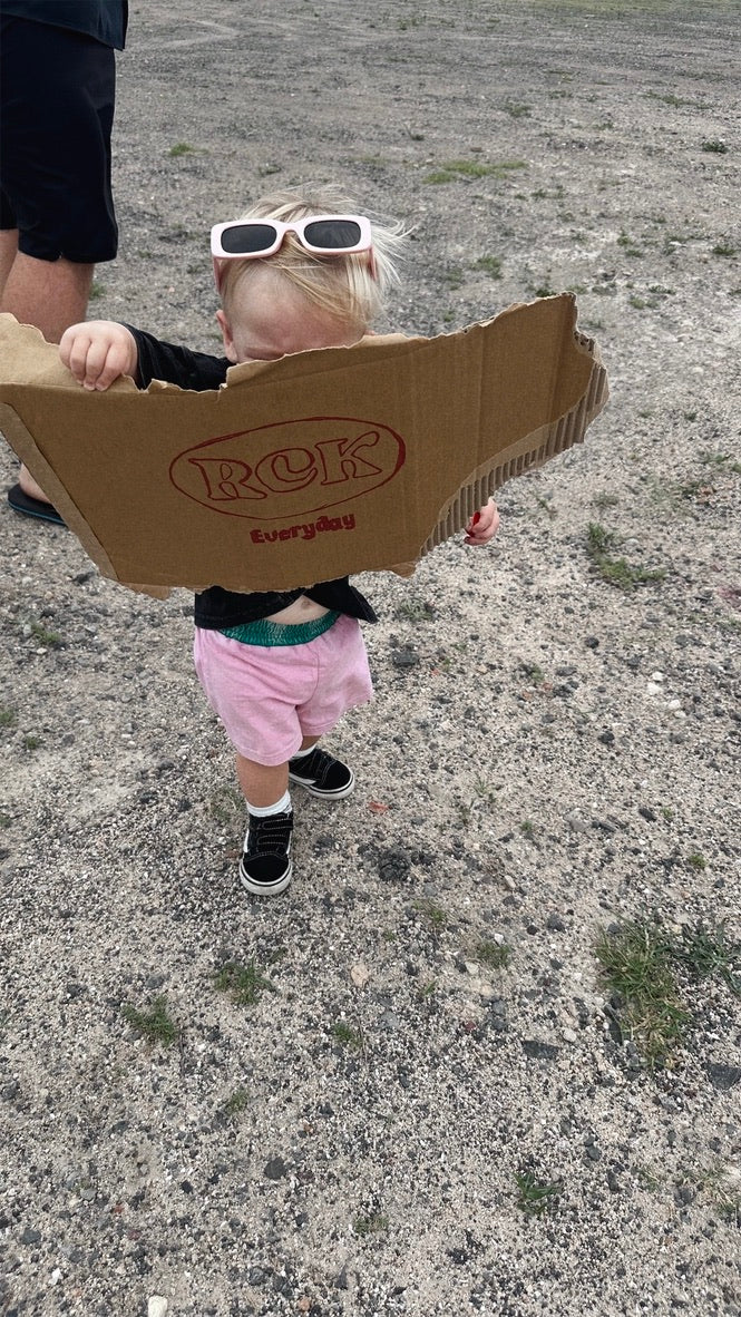 Child holding a cardboard box with 'Rock' branding outdoors on a gravelly surface.