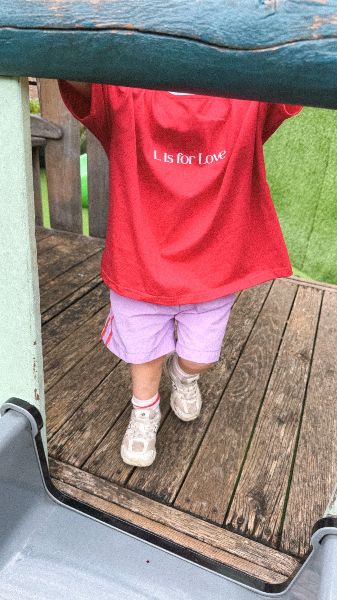 Child wearing a red shirt with 'L is for Love' text, standing on a wooden deck.