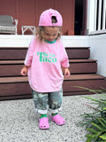 Child wearing a pink shirt with 'T is for Taco' text, pink cap, and pink shoes standing on a concrete surface.