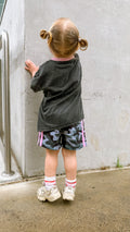 Child in a gray sweatshirt and camouflage shorts standing against a concrete wall.