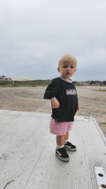 Child wearing a black t-shirt with pink text and pink shorts standing on a concrete surface.