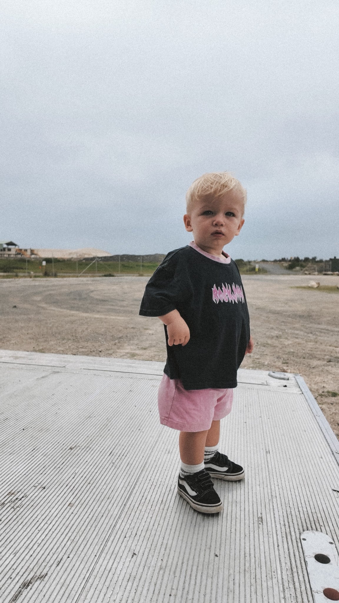 Child wearing a black t-shirt with pink text and pink shorts standing on a concrete surface.